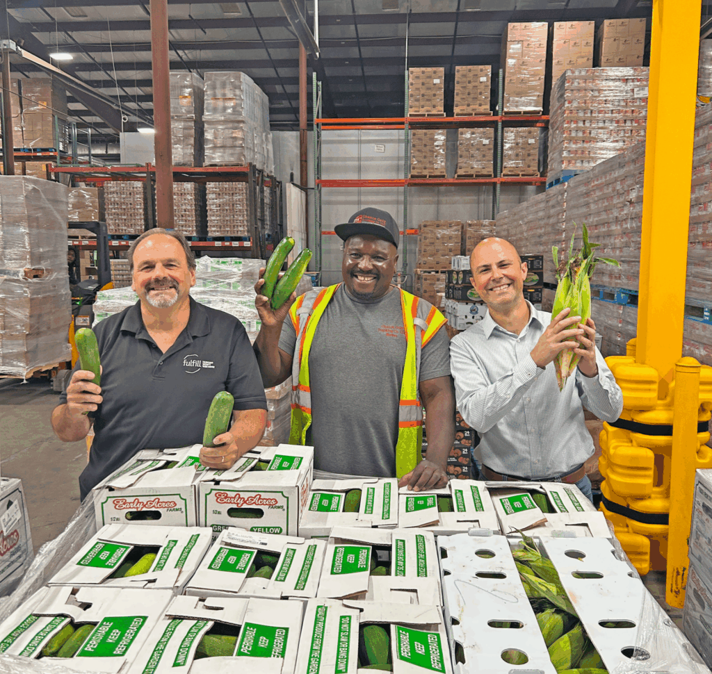 team members holding fresh zucchini, corn, and cucumbers in warehouse